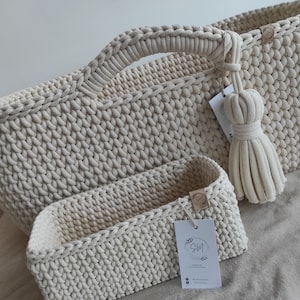 Puede incluir: Dos cestas de crochet de color crema, una ovalada con asa y otra rectangular más pequeña. La cesta ovalada tiene un borla colgando del asa. Ambas cestas tienen una pequeña etiqueta de madera con el texto "SM Handmade with love"
