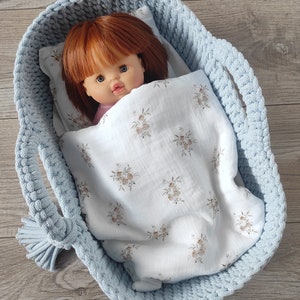 May include: A doll with red hair and blue eyes rests in a light blue crocheted basket. The doll is covered with a white blanket with a floral pattern. The basket has handles and a tassel.