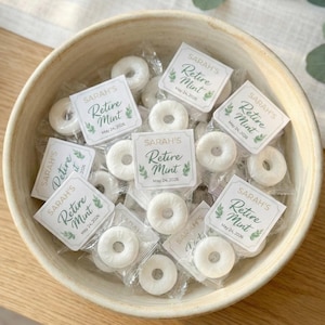 May include: A wooden bowl filled with individually wrapped white mint candies. Each candy wrapper has a label that reads "SARAH'S Retire Mint May 24, 2026" with a green leaf design. The bowl sits on a light-colored wooden surface.