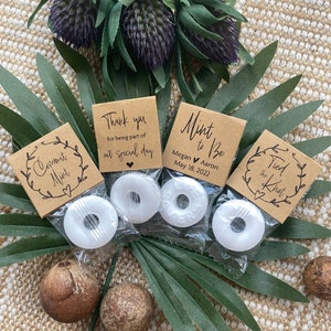 May include: Three small brown paper favor bags with white mints inside. The bags are decorated with handwritten text, including "Mint to Be", "Tied the Knot", and "Thank you for being part of our special day".