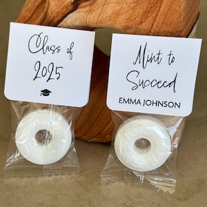 May include: Two small white bags with white mints inside. The bags have white labels with black text. One label says "Class of 2025" with a graduation cap icon. The other label says "Mint to Succeed" with the name "Emma Johnson" underneath.