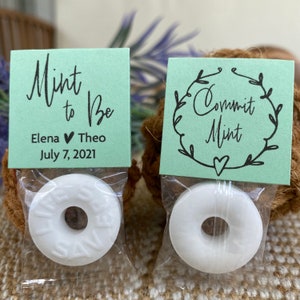 Puede incluir: Dos peque&ntilde;as bolsas de caramelos de menta con etiquetas verdes. Una etiqueta dice "Mint to Be" con los nombres Elena y Theo y la fecha 7 de julio de 2021. La otra etiqueta dice "Commit Mint" con una corona decorativa alrededor del texto.
