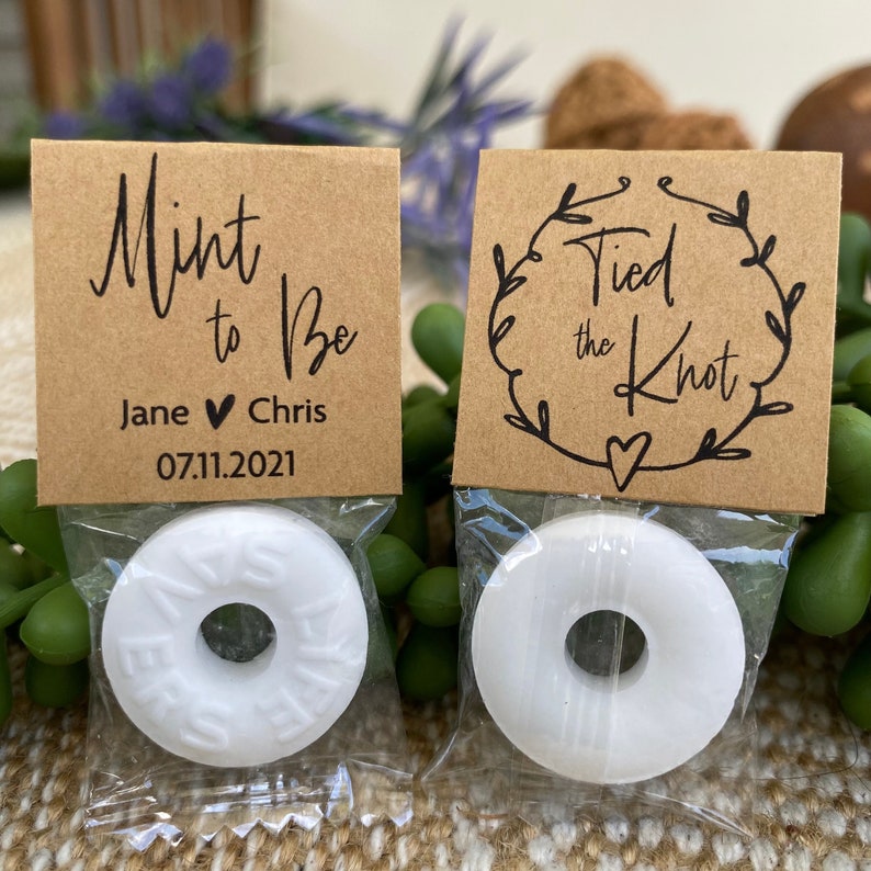Puede incluir: Dos peque&ntilde;as bolsas de papel marr&oacute;n con mentas blancas dentro. Las bolsas tienen texto. Una bolsa dice "Mint to Be" con los nombres Jane y Chris y la fecha 07.11.2021. La otra bolsa dice "Tied the Knot" con un dise&ntilde;o de corona.
