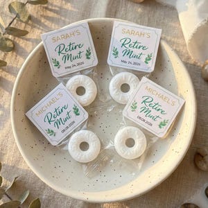May include: A ceramic bowl filled with individually wrapped white mints. Each mint is paired with a small card that reads "SARAH'S Retire Mint May 24, 2026" or "MICHAEL'S Retire Mint 09.08.2026" in green script. The bowl sits on a neutral-toned surface.