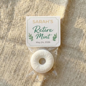 May include: A small cellophane bag containing a white mint candy with a hole in the center. The bag is attached to a small card that reads "SARAH'S Retire Mint" in green script, with the date "May 24, 2026" below.