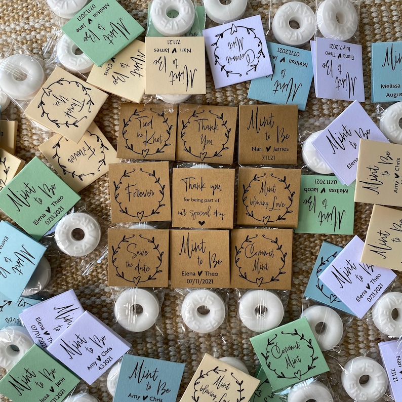 Puede incluir: Surtido de peque&ntilde;as mentas redondas blancas envueltas en bolsas de pl&aacute;stico transparentes con etiquetas de papel marr&oacute;n, verde y azul. Las etiquetas tienen varios textos, incluyendo "Mint to Be", "Save the Date", "Thank You" y "Commit Mint".