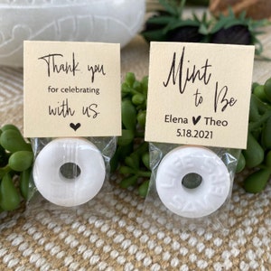 Puede incluir: Dos peque&ntilde;as bolsas blancas de regalo con una menta dentro y una etiqueta de agradecimiento. La etiqueta de la izquierda dice "Gracias por celebrar con nosotros" con un peque&ntilde;o coraz&oacute;n. La etiqueta de la derecha dice "Mint to be" con los nombres Elena y Theo y la fecha 5.18.2021.