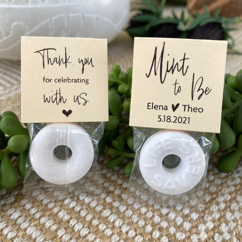 Puede incluir: Dos peque&ntilde;as bolsas blancas de regalo con una menta dentro y una etiqueta de agradecimiento. La etiqueta de la izquierda dice "Gracias por celebrar con nosotros" con un peque&ntilde;o coraz&oacute;n. La etiqueta de la derecha dice "Mint to be" con los nombres Elena y Theo y la fecha 5.18.2021.