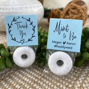 Puede incluir: Dos peque&ntilde;as bolsas de regalo azules con mentas blancas en su interior. Una bolsa tiene una etiqueta de agradecimiento con un dise&ntilde;o de corona. La otra bolsa tiene una etiqueta que dice "Mint to Be" con los nombres Megan y Aaron y el hashtag #freshlywed.