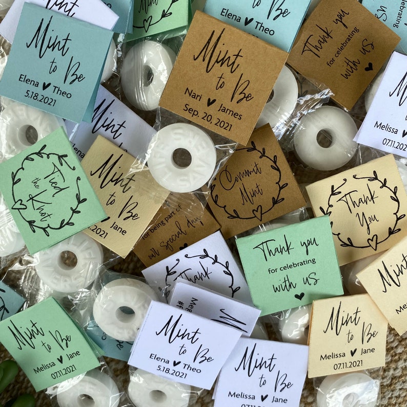 Puede incluir: Una colecci&oacute;n de peque&ntilde;as mentas redondas blancas envueltas en bolsas de pl&aacute;stico transparentes con varias etiquetas de papel marr&oacute;n, verde y blanco. Las etiquetas tienen texto escrito a mano que dice "Mint to Be" con diferentes nombres y fechas. Algunas etiquetas tambi&eacute;n tienen el texto "Gracias por celebrar con nosotros" o "Commit Mint".