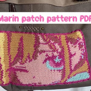 Könnte beinhalten: Ein handgefertigter Häkelaufnäher mit einem Charakterdesign in Rosa, Gelb und Blau. Der Aufnäher ist auf eine braune Canvas-Tasche genäht. Der Text über dem Aufnäher lautet "Marin patch pattern PDF".