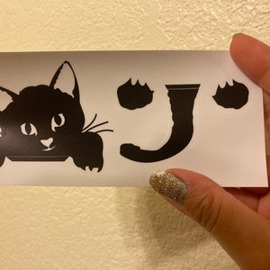 May include: Black cat peeking out from behind a white surface with the word "J" and paw prints on either side.