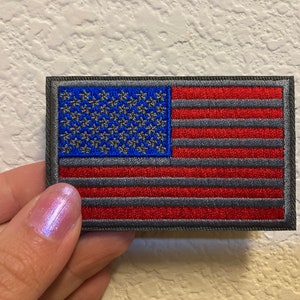 May include: A small embroidered American flag patch with a dark gray border and red and blue stitching. The stars are gray and the stripes are red and gray.