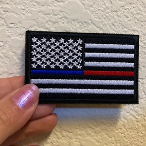 May include: A black embroidered patch with a thin blue line and a red line above the American flag. The flag has white stars on a blue field and white stripes on a black background.