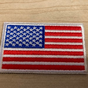 May include: A small embroidered American flag patch with white stars on a blue field and red and white stripes on a white background.