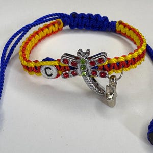 May include: A handmade bracelet with a blue, yellow, and red woven band. It features a silver dragonfly charm adorned with red and green rhinestones. The bracelet has blue adjustable cords and a small charm with the letter "C".