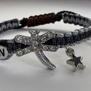 May include: Macrame bracelet in shades of gray and black, featuring a silver dragonfly charm with clear rhinestones, a star charm, and a circular charm with the letter "W". The bracelet is adjustable.