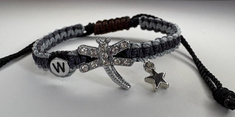 May include: Macrame bracelet in shades of gray and black, featuring a silver dragonfly charm with clear rhinestones, a star charm, and a circular charm with the letter "W". The bracelet is adjustable.