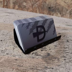 May include: A stack of gray business cards with a black and white logo of a stylized letter "D" with a diagonal line through it. The cards are in a black metal business card holder.