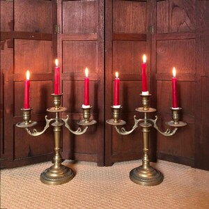 May include: Two matching brass candelabras, each featuring three arms and a central stem, hold a total of six lit red taper candles. The metallic candle holders have a polished finish with curved arms and circular bases, standing upright.