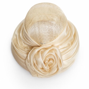 May include: A cream-coloured hat featuring a structured, woven dome crown and a wide, gathered satin band encircling the base. A large, multi-layered fabric rose ornament is attached to the front, matching the satin material of the band.