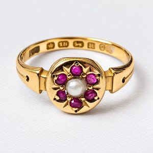 May include: A gold ring featuring a central white pearl surrounded by six small round-cut red rubies in a circular setting. The inner band is engraved with the number 18, a crown symbol, a lion passant, and the letter R, indicating gold purity.