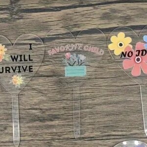Può includere: Cinque segnalibri per piante in acrilico a forma di cuore con diversi disegni floreali e testo. I segnalibri recitano "Favorite Child", "I Will Survive", "Favorite Child", "No Idea" e "Still Alive".