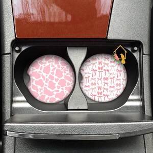 May include: Two car cup holders with pink and white patterned coasters. One coaster features a pink and white cow print pattern. The other coaster features a pink and white bow pattern.