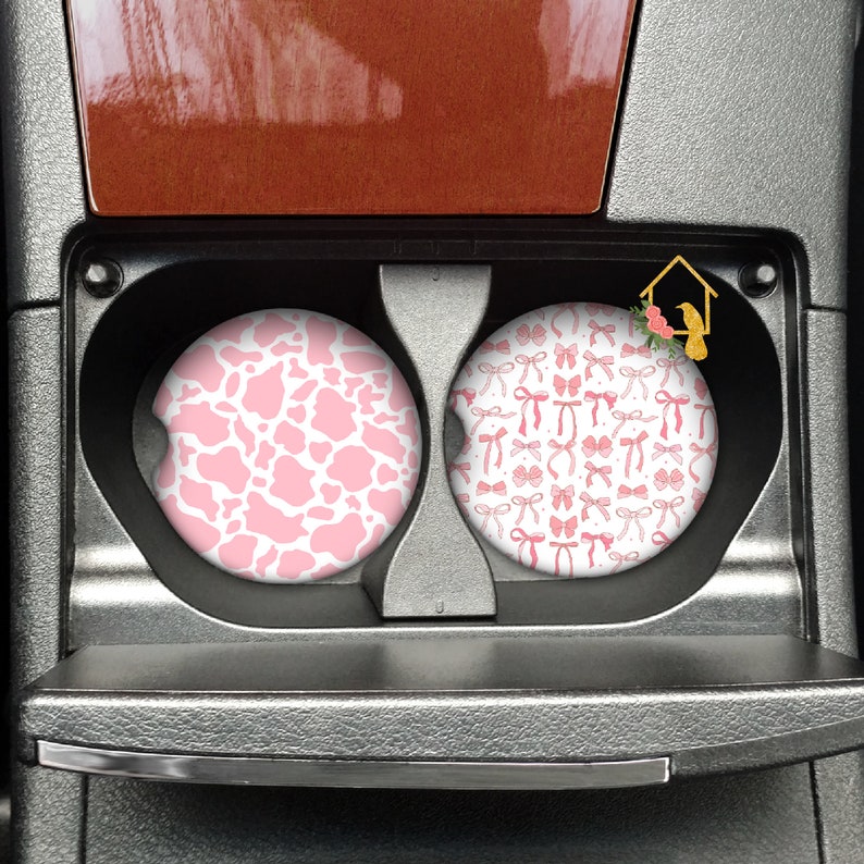 May include: Two car cup holders with pink and white patterned coasters. One coaster features a pink and white cow print pattern. The other coaster features a pink and white bow pattern.