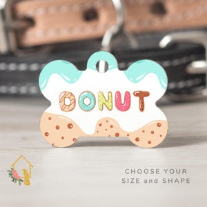 May include: A bone-shaped pet tag with a donut design. The tag features the word "DONUT" in colorful letters, with a light blue icing drip and a tan base with brown sprinkles. The tag has a hole for attaching to a pet collar.