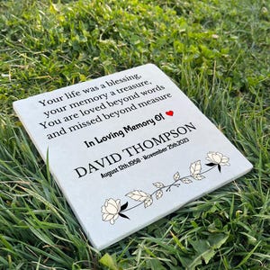 May include: A white memorial stone with black text that reads "Your life was a blessing, your memory a treasure, You are loved beyond words and missed beyond measure. In Loving Memory Of DAVID THOMPSON August 12th, 1958 - November 25th, 2023". The stone has a floral design with white flowers and green leaves.