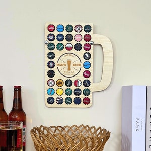 Può includere: Decorazione da parete in legno a forma di boccale di birra con una cornice in legno marrone e un manico ritagliato. Il boccale è decorato con tappi di bottiglia di vari colori e motivi. Il testo "Dad's Beer" è stampato sul boccale.