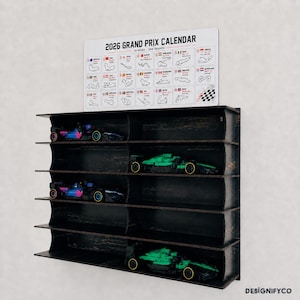 May include: A black wooden wall-mounted display shelf with five tiers, holding four toy race cars. Above the shelf is a white sign titled 2026 GRAND PRIX CALENDAR, featuring a grid of 23 race track outlines, country flags, and event dates.