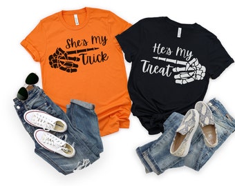 May include: Two t-shirts with a skeleton hand graphic and text. The orange shirt says "She's My Trick" and the black shirt says "He's My Treat".