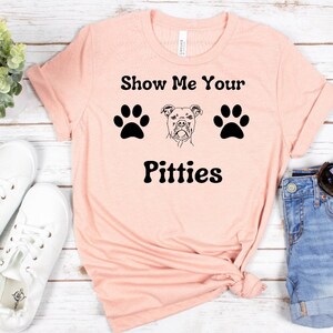 以下が含まれることがあります： 「Show Me Your Pitties」の黒い文字、足跡、犬のイラストが描かれた、薄いピンク色のTシャツ。 Tシャツは折りたたまれており、背景には白いスニーカーとデニムショーツがあります。