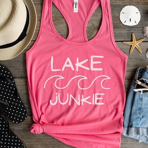 May include: A pink tank top with the words "LAKE JUNKIE" in white, with wave graphics. The image also includes a straw hat, black and white polka dot flip-flops, denim shorts, sunglasses, seashells, and a starfish, all arranged on a wooden surface.