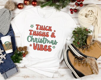 Puede incluir: Camiseta blanca con el texto "Thick Thighs & Christmas Vibes" en letras rojas y verdes con estrellas sobre un fondo blanco.