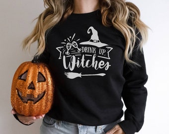 Op de afbeelding: Zwart sweatshirt met een wit grafisch ontwerp van een heksenhoed, twee champagne glazen en de tekst "Drink Up, Witches".