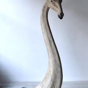 May include: A wooden sculpture of a giraffe's head and neck, with a long, curved neck and a detailed head. The sculpture is painted in a light gray color and is standing on a black base.