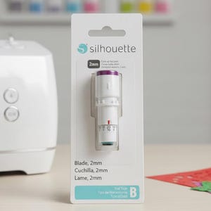 May include: A white Silhouette cutting blade, 2mm, in its original packaging, with the Silhouette logo. The packaging also lists the blade's specifications in English and Spanish. A white cutting machine is visible in the background, along with colorful cutouts.