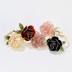 May include: Five floral napkin rings. Each ring features a different colored rose: cream, burgundy, pink, lavender, and navy blue. The roses have gold wire detailing and green leaves. The rings are made of metal.