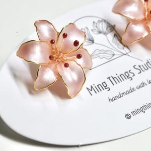 Puede incluir: Un par de pendientes de flores rosas con detalles de alambre dorado y centros rojos. Los pendientes est&aacute;n hechos a mano y presentan un delicado dise&ntilde;o floral.