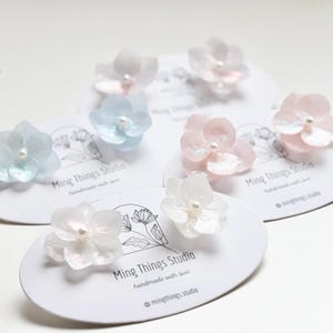 Könnte beinhalten: Vier Paare zarte, blumenförmige Ohrringe in Weiß, Hellblau und Rosa. Die Ohrringe sind handgefertigt und haben einen perlmuttartigen Mittelpunkt. Die Ohrringe werden auf weißen Karten mit dem Text "Ming Things Studio" und "handgefertigt mit Liebe" präsentiert.