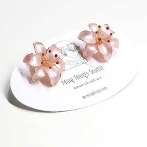 Puede incluir: Un par de pendientes de flor de lirio rosa con alambre dorado y cuentas rojas. Los pendientes est&aacute;n sobre una tarjeta blanca con el texto "Ming Things Studio handmade with love @mingthings.com"