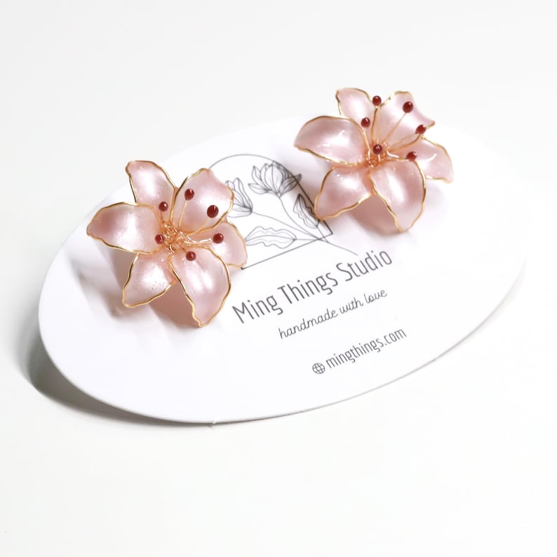Puede incluir: Un par de pendientes de flor de lirio rosa con alambre dorado y cuentas rojas. Los pendientes est&aacute;n sobre una tarjeta blanca con el texto "Ming Things Studio handmade with love @mingthings.com"