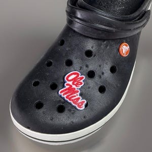 May include: Black clog shoe with a white sole stripe. The shoe features a red, white, and blue "Ole Miss" patch and a round orange logo charm. The shoe has multiple ventilation holes.