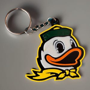 May include: A keychain featuring a cartoon duck mascot with a green hat, orange beak, and yellow bow tie. The duck is outlined in black and white, with a silver key ring and chain attached. The keychain is a vibrant accessory.
