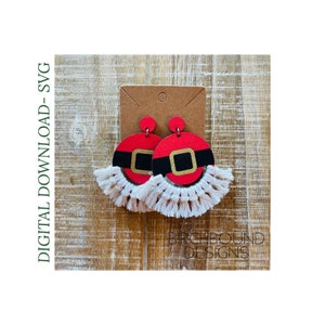 May include: A pair of red and black Santa Claus-themed earrings with white fringe. The earrings are made of wood and have a gold buckle detail. The earrings are displayed on a brown wooden background.