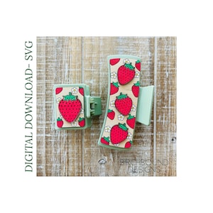 Può includere: Due mollette per capelli verdi con un pannello di legno con un motivo a fragole. Il pannello di legno è decorato con fragole rosse e fiori bianchi. Il testo "BIRCHBOUND DESIGNS" è visibile nell'angolo in basso a destra dell'immagine.