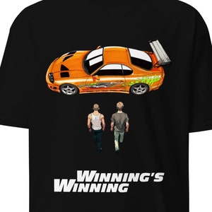 May include: Black t-shirt featuring an orange sports car graphic with green and white accents, and two figures walking away. The text "Winning's Winning" is displayed in white at the bottom of the shirt.